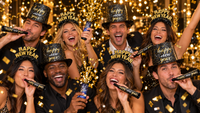 Friends celebrating New Year's Eve in black and gold Happy New Year top hats and glitter tiaras from the Confetti New Year Party Kit, blowing printed horns and popping confetti bursts